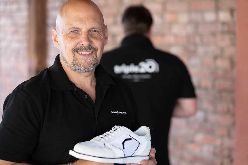 Triple20 Dartsshoes Gründer Jürgen Bloch hält einen triple20 Sneaker in der Hand, im Hintergrund sieht man verschwommen seinen Partner Wolfgang Marx
