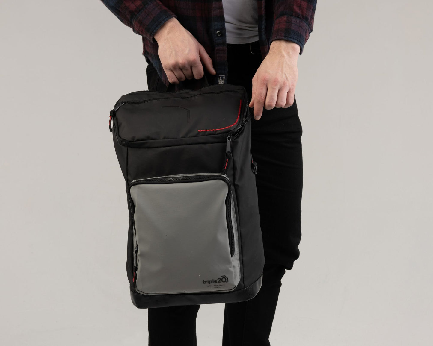 Ein männliches Model hält den neuen triple20 Dartrucksack in den Händen. Gut zu sehen ist der Rucksack in seiner Gesamtheit sowie die lässige Farbkombi aus schwarz, grau und rot. Der Rucksack hat eine standardisierte Größe und fasst neben einem Paar Schuhe auch diverse weitere Utensilien.